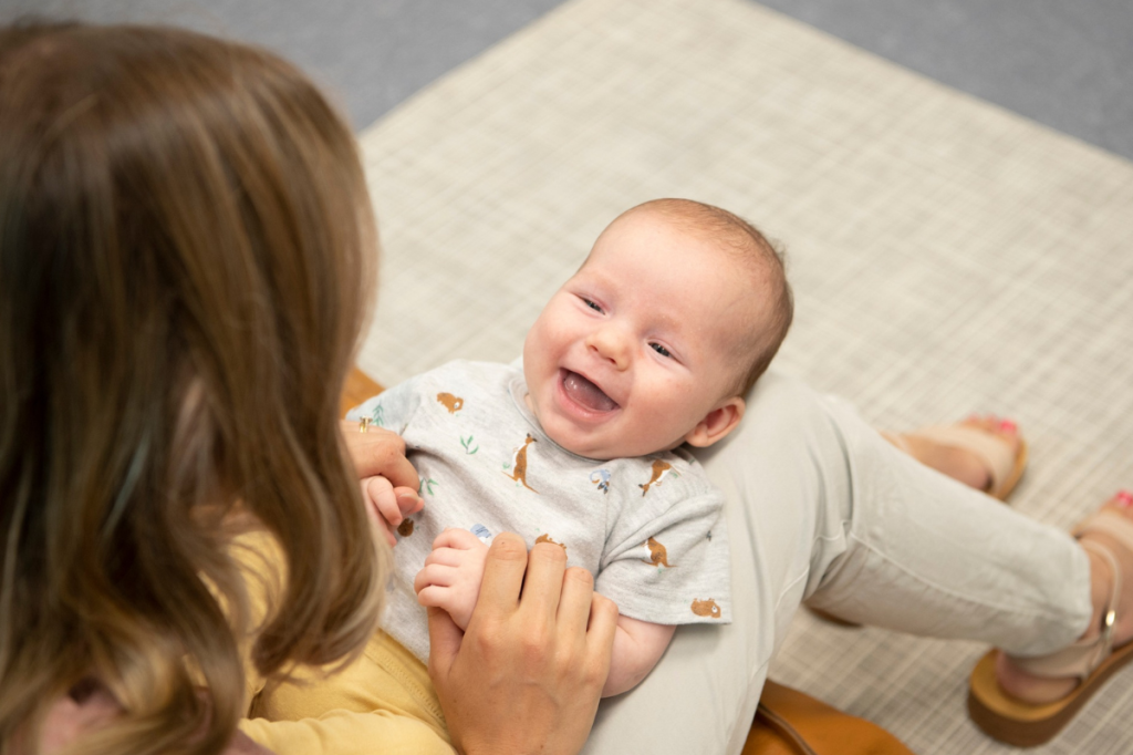 baby sitting on mother’s lap holding her hands and looking up at her while laughing