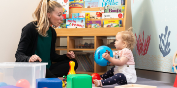 Adult playing with baby on floor as baby holds a ball and looks at them