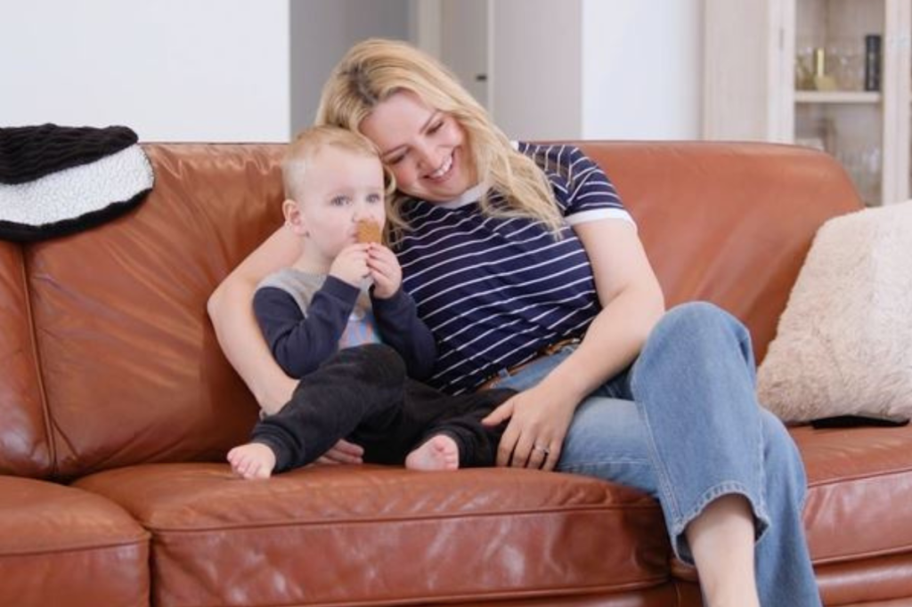 mother smiling and cuddling baby on couch while baby holds a biscuit and looks ahead