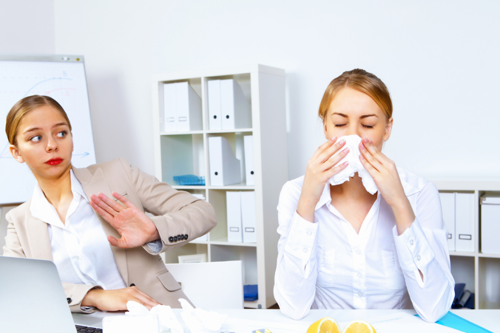Sick woman blowing her nose at work while her colleague tries to move further away from her.