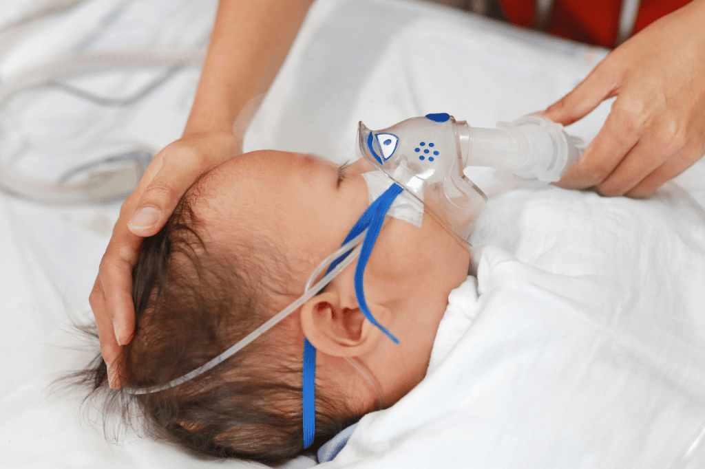 baby wearing oxygen mask while lying in hospital bed