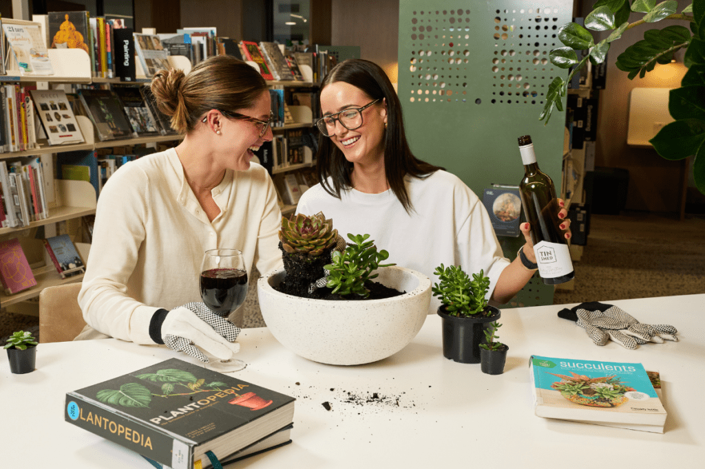 Two young women enjoying a glass of wine while they pot succulents in a South Australian library.